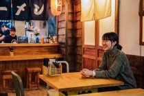 Задоволена жінка в азіатському светрі, дивлячись з зубатою посмішкою, сидячи за дерев'яним столом у барі. — стокове фото