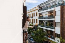 Vue latérale d'une jeune femme noire admirant des façades et des arbres de bâtiments urbains à plusieurs étages, debout sur un balcon clôturé et regardant loin — Photo de stock