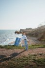 Vista laterale di positivo giovane artista donna in abito elegante e cappello a piedi vicino alla spiaggia sabbiosa del mare ondulato con pittura in mano — Foto stock
