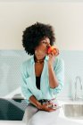 Belle femme afro-américaine moderne avec smartphone à la main assis sur le comptoir de la cuisine regardant loin à la maison et mangeant de la pomme — Photo de stock