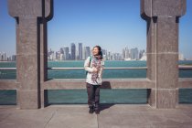Vue de face du joyeux touriste asiatique souriant à la caméra tout en restant seul à côté de la clôture cintrée sur le front de mer dans le Musée d'Art islamique contre West Bay et vue magnifique sur le centre-ville de Doha — Photo de stock