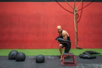 Potente deportista con bolsa pesada pisando y abalanzándose sobre el taburete durante el entrenamiento funcional en el gimnasio moderno - foto de stock