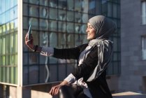 Vista lateral de una mujer musulmana positiva en hijab tomando autorretrato en un teléfono inteligente en la ciudad en un día soleado - foto de stock