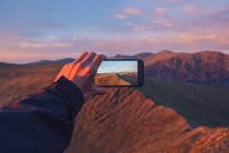 Caminante masculino anónimo de cultivos tomando fotos de las tierras altas en el teléfono inteligente al atardecer en Gales - foto de stock