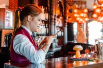 Vista lateral de la alegre camarera femenina tomando fotos de cóctel agrio en vidrio servido en el mostrador en el pub - foto de stock