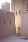 Полная длина радостный азиатский путешественница смотрит в сторону и улыбается во время прогулки по улице против бежевого и коричневого фасадов старых зданий с арочными окнами в Дохе — стоковое фото