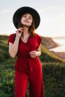 Menina jovem atraente em sundress vermelho e chapéu em pé no prado gramado verdejante no campo ensolarado — Fotografia de Stock