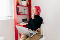 Freelancer jovem focado em roupas casuais sentadas na cadeira e usando laptop enquanto trabalhava no projeto em apartamento moderno leve — Fotografia de Stock