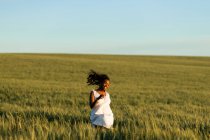 Lächelnde junge schwarze Dame in weißem Sommerkleid schlendert auf grünem Weizenfeld und schaut tagsüber unter blauem Himmel weg — Stockfoto