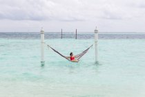 Woman in red swimsuit lying in hammock swing over ocean surf line relaxing in Maldives on cloudy day — Stock Photo