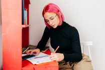 Vista lateral de la joven diseñadora creativa con pelo rosa en traje casual y gafas usando smartphone y boceto de dibujo mientras trabaja en el escritorio en casa - foto de stock