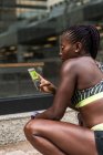 Femme afro-américaine en vêtements de sport naviguant smartphone moderne tout en se tenant debout sur fond flou de la rue de la ville pendant l'entraînement en plein air — Photo de stock