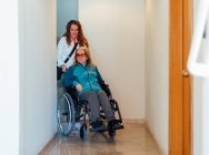 Взрослая женщина, толкающая инвалидное кресло со старшей матерью и идущая по коридору в реабилитационном центре — стоковое фото