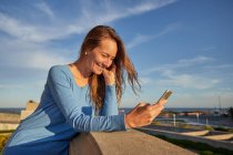 Sonriendo señora adulta navegando por teléfono mientras se apoya en la cerca cerca del océano en la calle de la ciudad en el día soleado - foto de stock