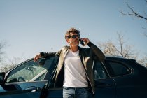 Male in sunglasses talking on cellphone while standing near automobile on sunny day — Stock Photo