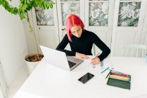 From above of thoughtful female freelance designer in casual outfit and glasses working at table with laptop and drawing fashion sketches with pencils — Stock Photo