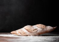 Baguette appétissante fraîchement cuite avec croûte croustillante placée sur une table en bois recouverte de farine blanche sur fond noir — Photo de stock