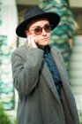 Вид сбоку андрогинного человека в шляпе и современных солнцезащитных очках, говорящего по мобильному телефону, глядя на камеру, стоящую на улице при дневном свете — стоковое фото