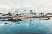 Mar frío con aguas tranquilas cerca de asentamientos costeros y cumbres nevadas en un día nublado de invierno en las Islas Lofoten, Noruega - foto de stock