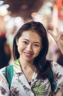 Happy Asian female traveler in tropical shirt with backpack looking away while standing on bazaar against blurred crowd in Doha — Stock Photo