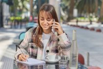 Positivo giovane donna prendere appunti in notebook durante la conversazione telefonica mentre seduto a tavola in caffè all'aperto in città — Foto stock