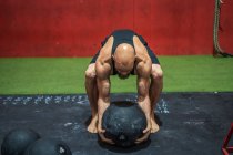 Сильный спортсмен поднимает тяжелый мяч с пола во время тренировки в современном спортзале — стоковое фото