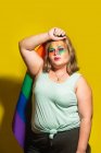 Mulher com excesso de peso com maquiagem criativa segurando bandeira do arco-íris e cabeça tocando contra fundo amarelo — Fotografia de Stock