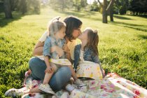 Симпатичні маленькі сестри цілують щасливу матір, відпочиваючи разом на ковдрі на зеленому газоні в літньому парку — стокове фото