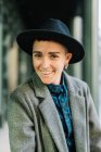 Cheerful young transgender person in classy coat and hat looking at camera in daylight — Stock Photo