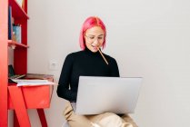 Очаровательная девушка-фрилансер в повседневной одежде, сидящая на стуле и использующая ноутбук во время работы над проектом в светлой современной квартире — стоковое фото