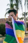 Tranquilo hombre gay con los ojos cerrados envueltos en colorida bandera LGBT en la calle de la ciudad - foto de stock