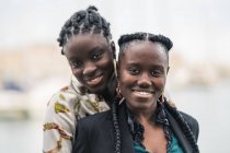 Zufriedene Afroamerikanerinnen, die in der Nähe bleiben und bei hellem Tag mit nachdenklichem Lächeln in die Kamera schauen — Stockfoto