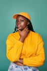 Hembra afroamericana regordeta sin emociones en suéter amarillo y gorra tocando la barbilla y mirando a la cámara mientras está de pie contra la pared azul - foto de stock