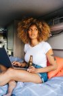 Young African American female traveler looking away with curly hair drinking beer and watching movie on laptop while resting inside camper van during summer holidays — Stock Photo