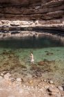 Relaxado mulher asiática olhando para trás para a câmera em água transparente de Bimmah Sinkhole cercado por rochas ásperas durante a viagem em Omã — Fotografia de Stock
