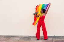 Femme afro-américaine joyeuse dans des vêtements élégants avec bouteille de boisson alcoolisée et drapeau coloré regardant loin sur fond clair — Photo de stock