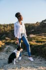 Full body side view of happy ethnic woman with Border Collie dog walking together on trail among grassy hills in sunny spring evening — Stock Photo