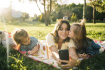 Щаслива молода жінка і милі маленькі дочки лежать на ковдрі і приймають селфі на смартфон, розважаючись разом на зеленому лузі в літньому парку — стокове фото