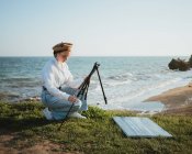 Seitenansicht der jungen stilvollen Malerin in lässiger Kleidung und Baskenmütze Aufstellen Staffelei auf grasbewachsenem Gelände über Sandstrand des welligen Ozeans an sonnigen Tag — Stockfoto