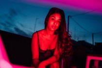 Encantada joven hispana de etnia femenina en lencería mirando a la cámara mientras descansa en la terraza bajo la luz roja de neón en la noche con el cielo azul oscuro - foto de stock