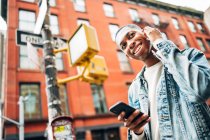 Знизу веселий афроамериканець у джинсовій куртці серфінг сучасні мобільні телефони під час прогулянок по місту. — стокове фото