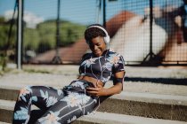 Молода афроамериканська жінка в барвистому спортивному вбранні слухає музику в навушниках, сидячи на кам 