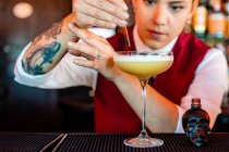 Profissional jovem garçom do sexo feminino adicionando álcool de garrafa em forma de crânio com conta-gotas em vidro enquanto prepara coquetel azedo no bar — Fotografia de Stock