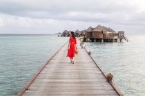 Rückansicht einer Frau im roten Kleid, die barfuß auf der tropischen Strandpromenade der Malediven entspannen will — Stockfoto