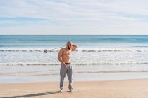 Full length of adult sportive man looking away doing around body pass with weight plate on band while warming up on sunny coast — Stock Photo