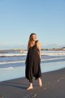 Улыбающаяся женщина в летнем платье стоит на песчаном берегу и смотрит в камеру — стоковое фото