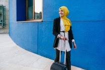 Cheerful Muslim female in headscarf standing with luggage in street against blue wall — Stock Photo