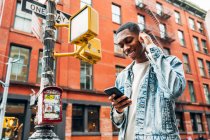 Знизу веселий афроамериканець у джинсовій куртці серфінг сучасні мобільні телефони під час прогулянок по місту. — стокове фото