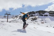 Atleta masculino anônimo em máscara de tecido pulando com snowboard sobre a neve contra a Serra Nevada e via cabo na Espanha — Fotografia de Stock