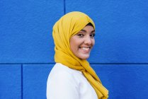 Вид сбоку на модную мусульманку в жёлтом хиджабе, стоящую, смотрящую на камеру на улице на синем фоне — стоковое фото